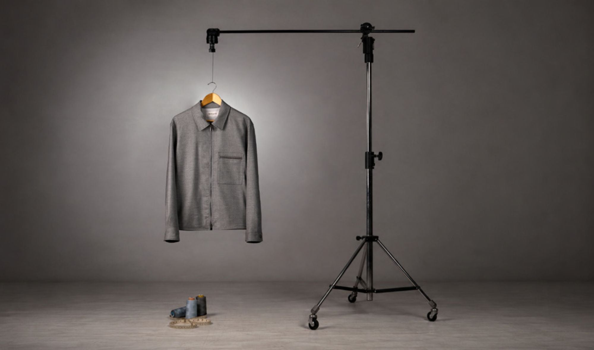Camicia grigia elegante appesa a una struttura di supporto per fotografia, con fondo semplice grigio e due matasse di filo poste sul pavimento in primo piano.