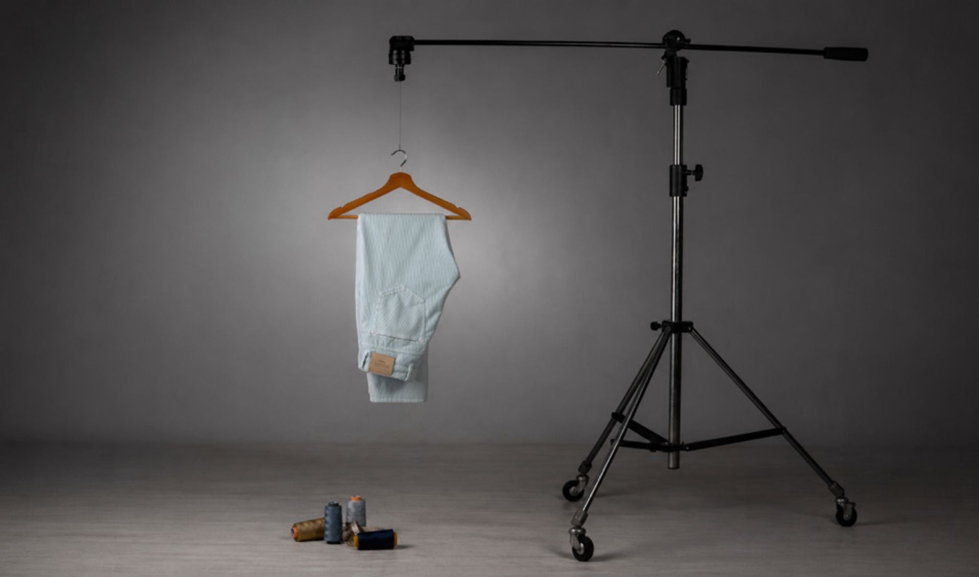 Pantaloni in denim chiaro appesi a un supporto da studio fotografico con sfondo grigio neutro, insieme a fili e accessori per il cucito posizionati a terra davanti.