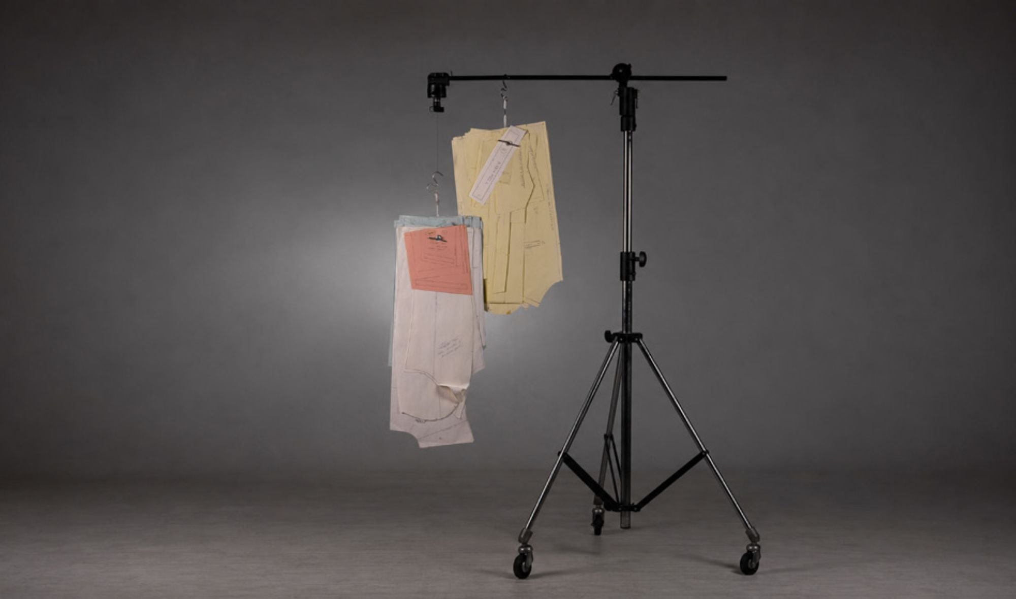 Cavalletto nero con ruote che sostiene due appendini con pantaloni chiari appesi, sfondo grigio uniforme e illuminazione soffusa su abbigliamento sartoriale.