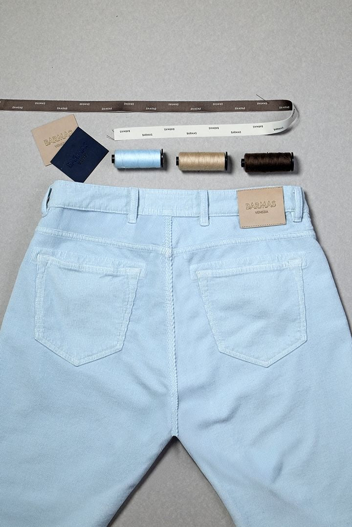 Dettaglio della parte posteriore di un paio di pantaloni in denim chiaro con etichetta in pelle Barmas, affiancati da tre rocchetti di filo colorato e strisce campione con nomi dei tessuti, su sfondo neutro.