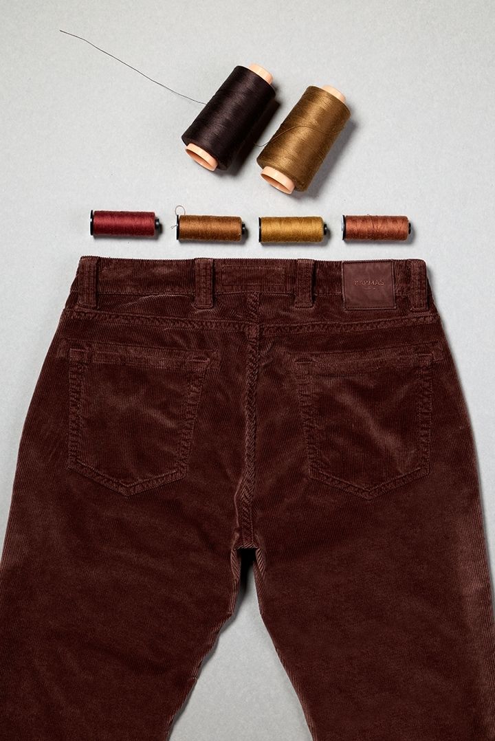 Pantaloni in velluto bordeaux disposti con cinque rocchetti di filo da cucito in tonalità calde sopra, rappresentando sartorialità e attenzione ai dettagli artigianali.