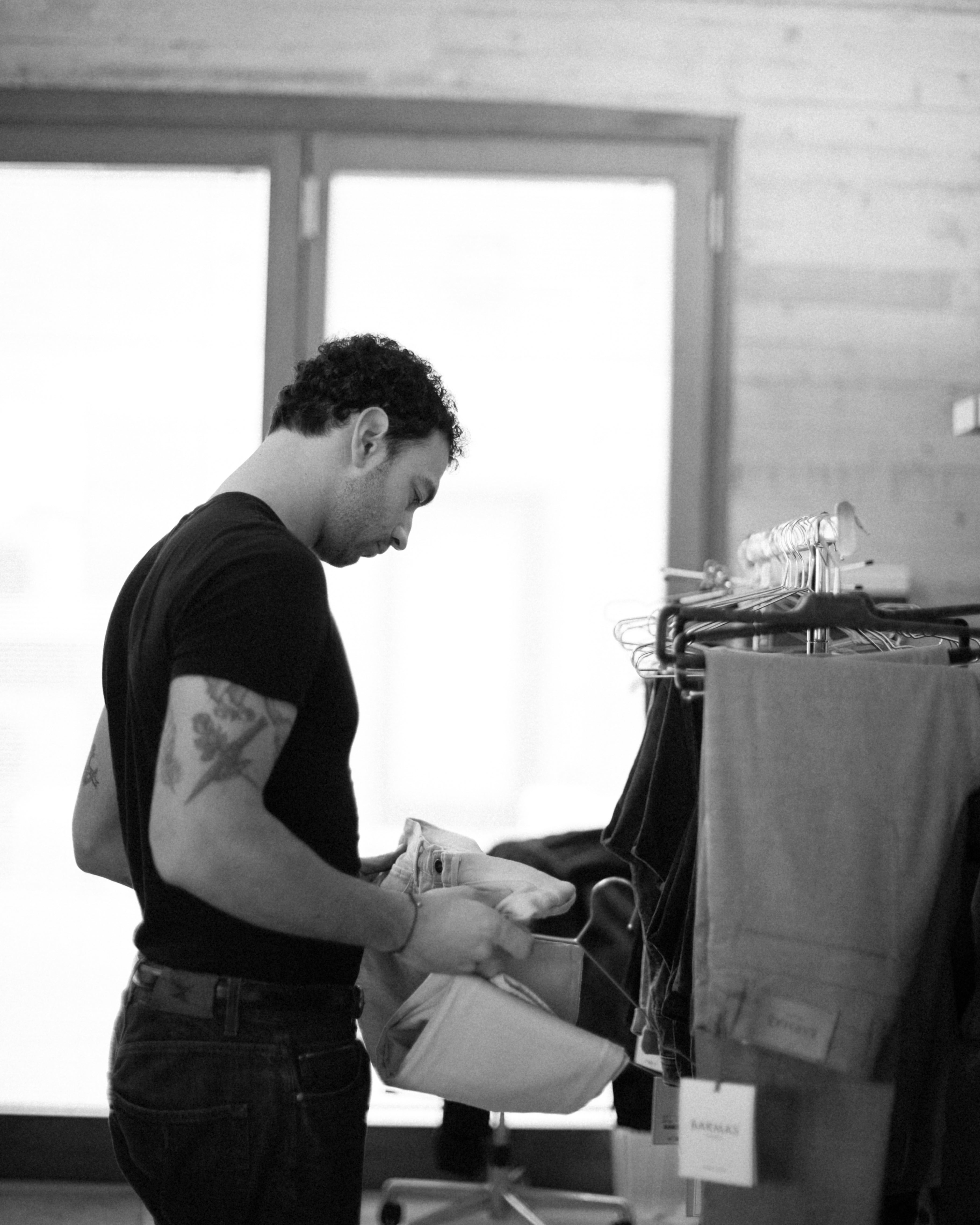 Uomo con tatuaggi in maglietta nera che osserva un paio di jeans in un atelier, fotografato in bianco e nero con atmosfera intima e concentrata.