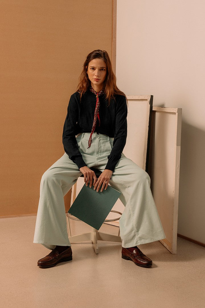 Donna seduta con pantaloni ampi chiari, camicia nera e foulard sottile, che tiene una cartella verde, sfondo semplice con toni caldi neutri.
