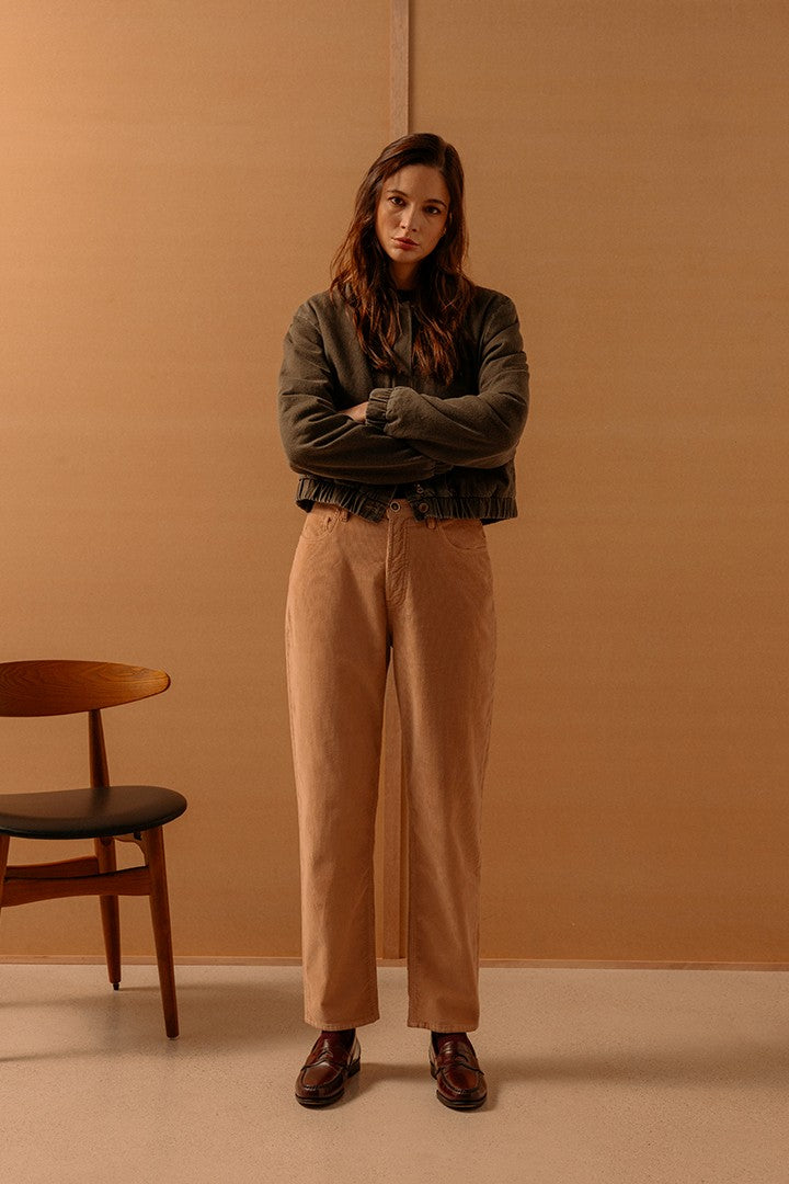 Donna in posa con braccia incrociate, indossa giacca verde scuro e pantaloni beige dritti, abbinati a scarpe marroni, su sfondo semplice color legno con sedia accanto.