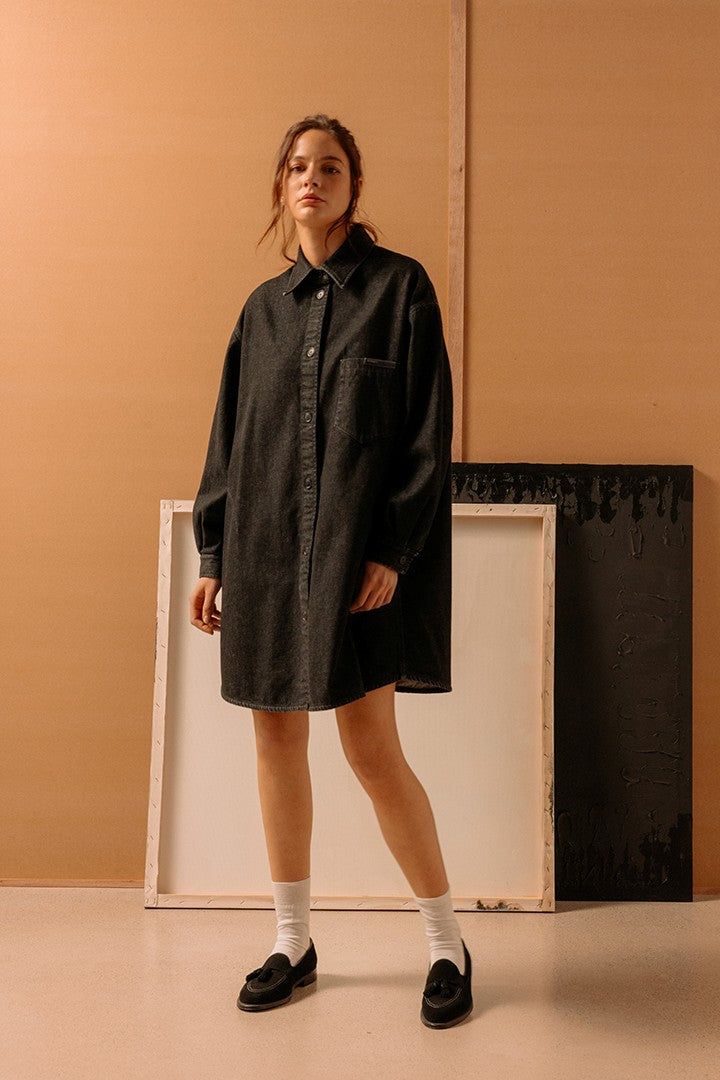 Donna in piedi con abito camicia oversize nero, calzini bianchi e mocassini neri, sfondo con cornici e parete beige in stile minimalista.