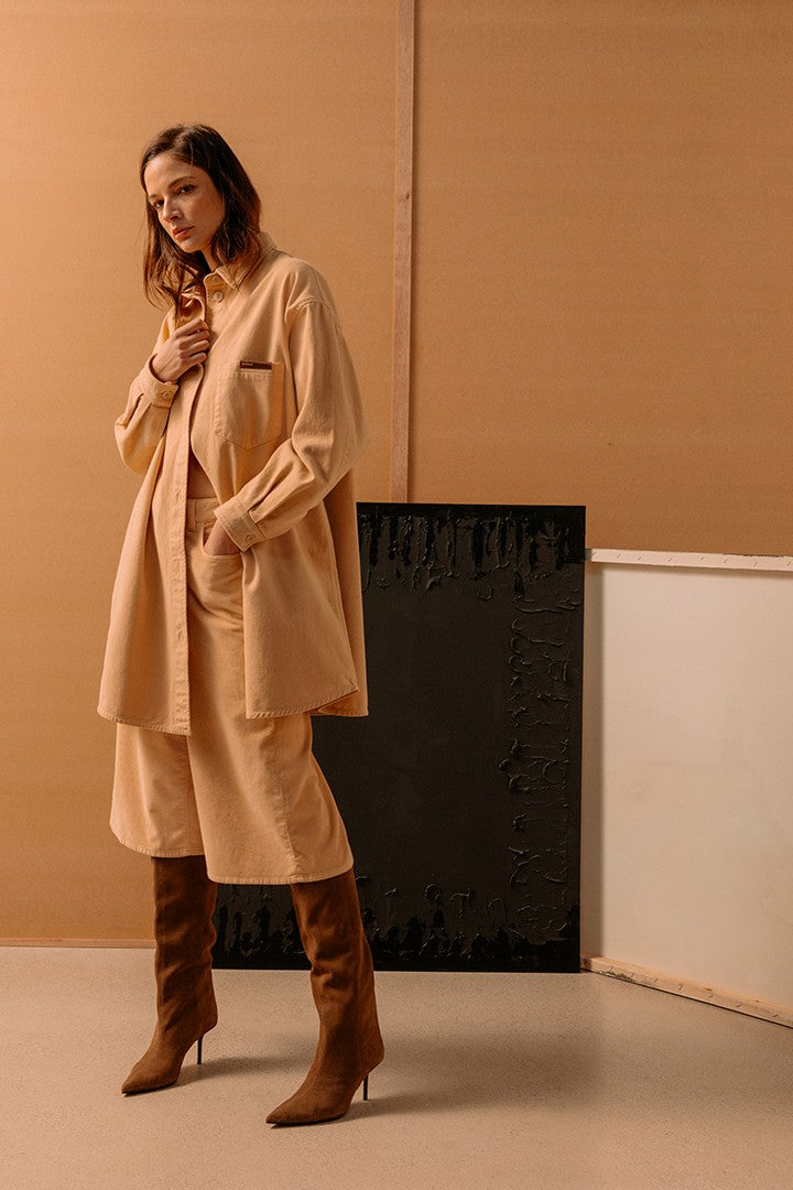 Donna in posa con outfit beige composto da camicia oversize, pantaloni larghi e stivali alti in pelle marrone, sfondo minimalista con pannelli beige e nero.