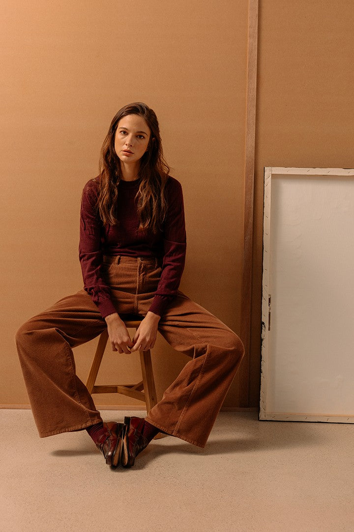 Donna seduta con pantaloni in velluto marrone ampio, maglione bordeaux e scarpe abbinate, su sfondo beige minimalista con cornice bianca appoggiata al muro.