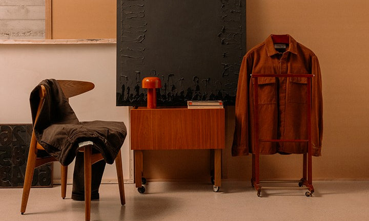 Ambiente interno minimalista con sedia di legno scuro coperta da capo in tessuto, mobiletto in legno con libri e lampada rossa, giacca marrone appesa su supporto con ruote, sfondo neutro.