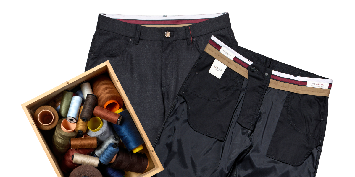 Due pantaloni in tessuto elegante, uno mostrato fronte e l'altro al rovescio per evidenziare la fodera interna, accanto a una scatola di fili da cucito di vari colori.