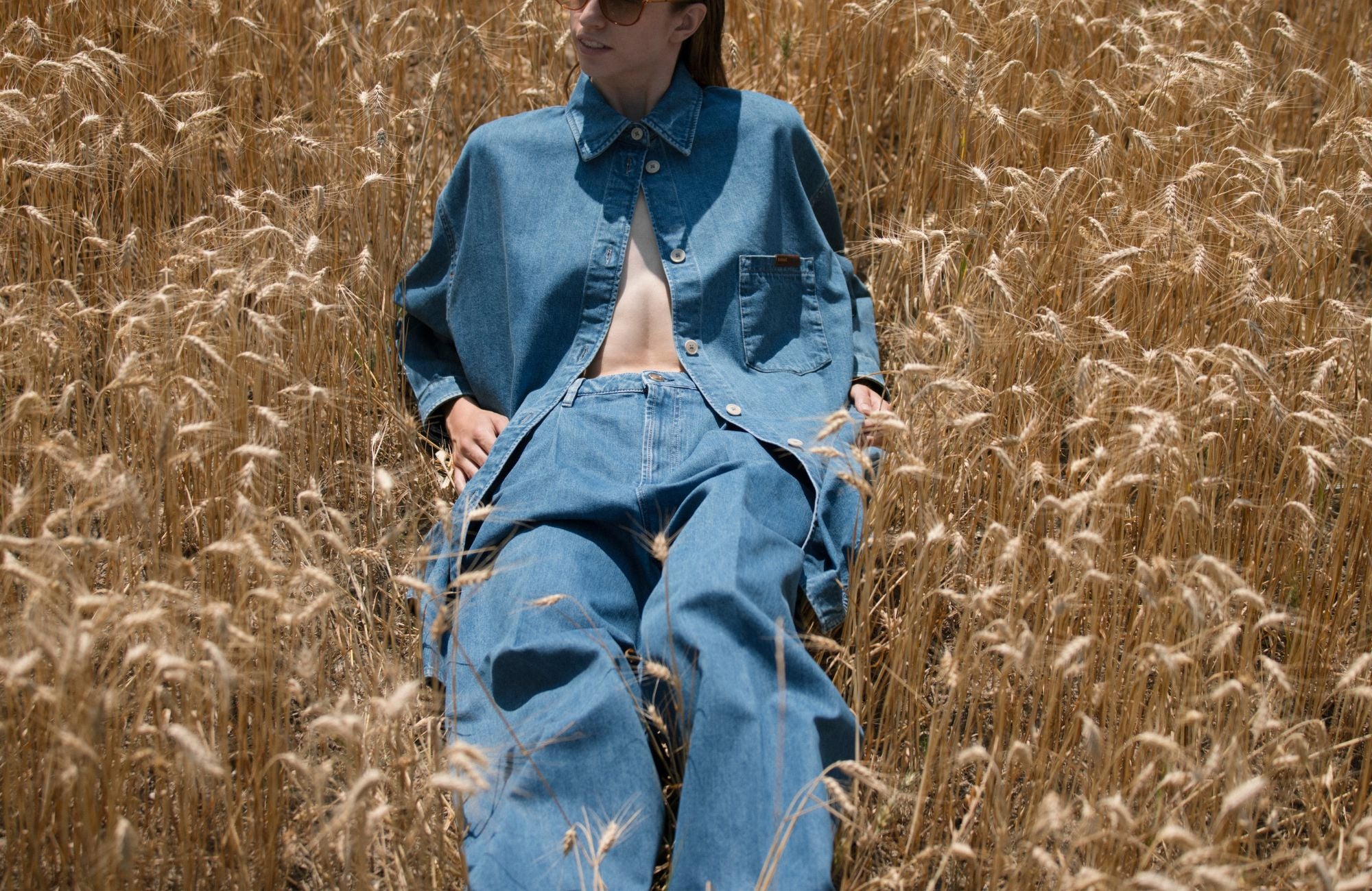 Donna con completo in denim blu sdraiata in un campo di grano, collezione SS26 Barmas