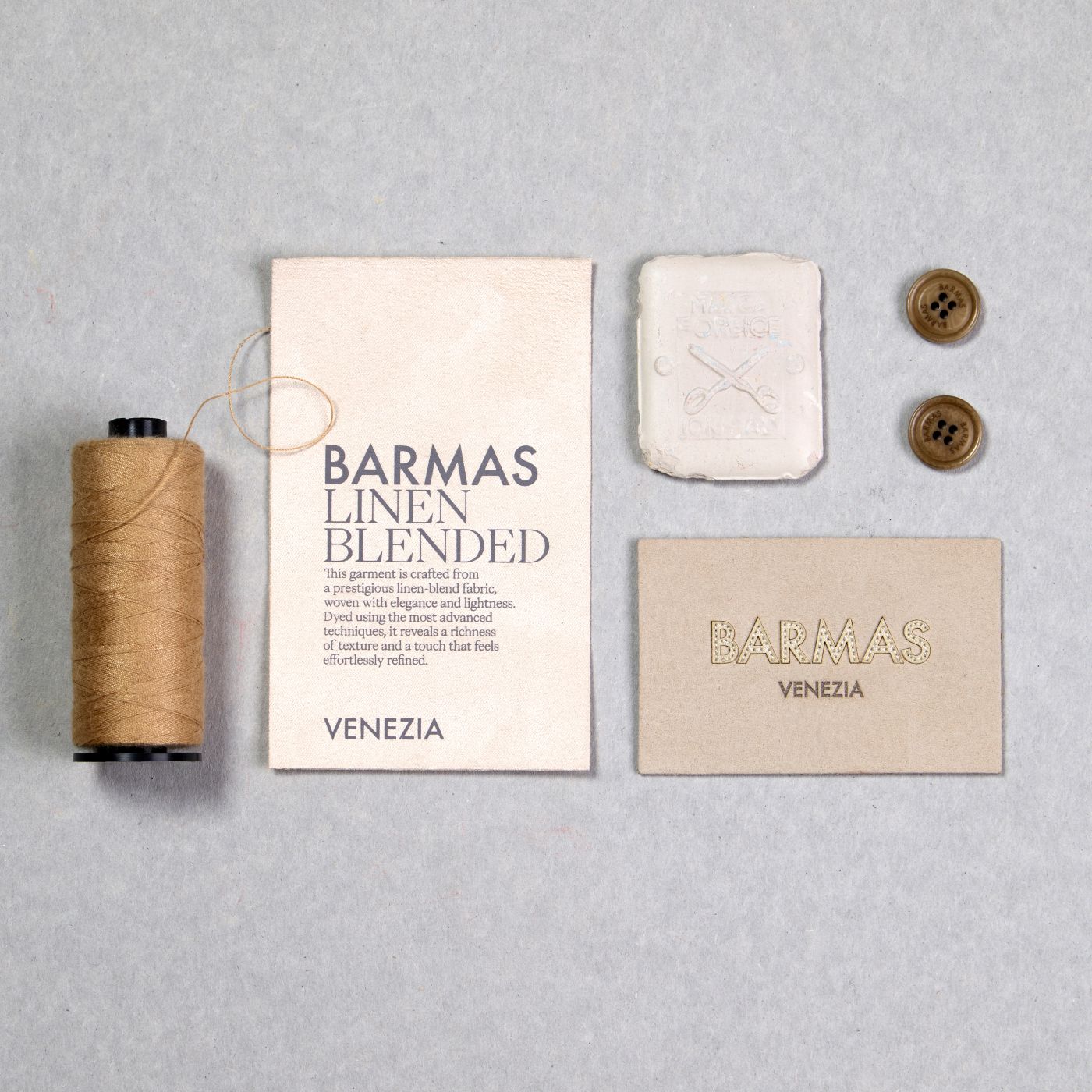 Materiali sartoriali Barmas con etichette, filo, bottoni e sapone da laboratorio su fondo neutro.