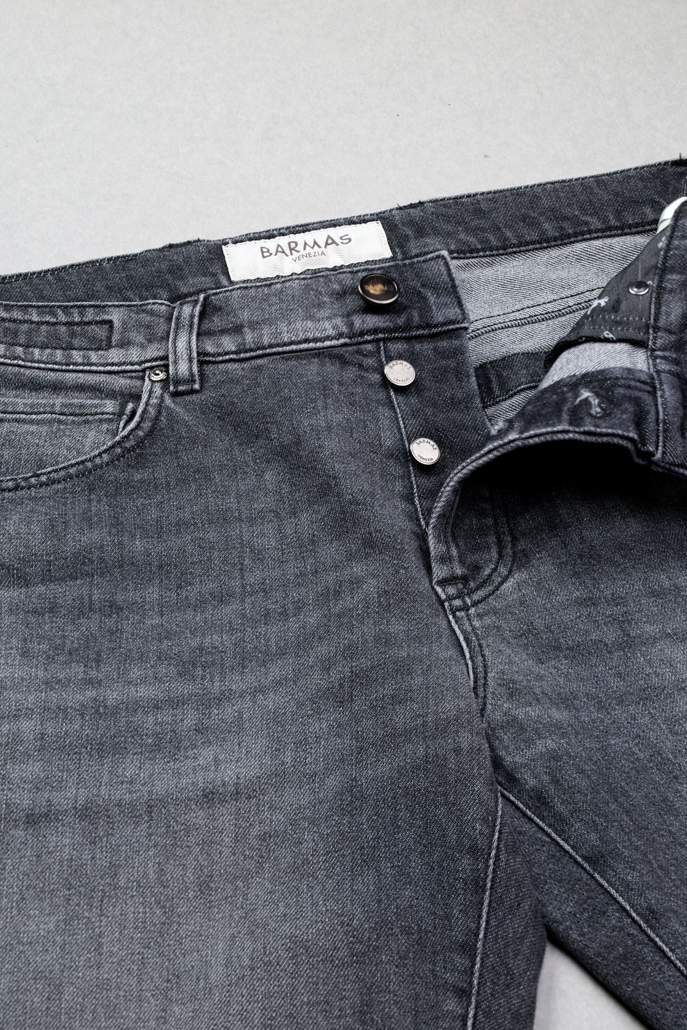 Dettaglio di jeans grigio scuro Barmas con bottoni metallici e costruzione sartoriale.