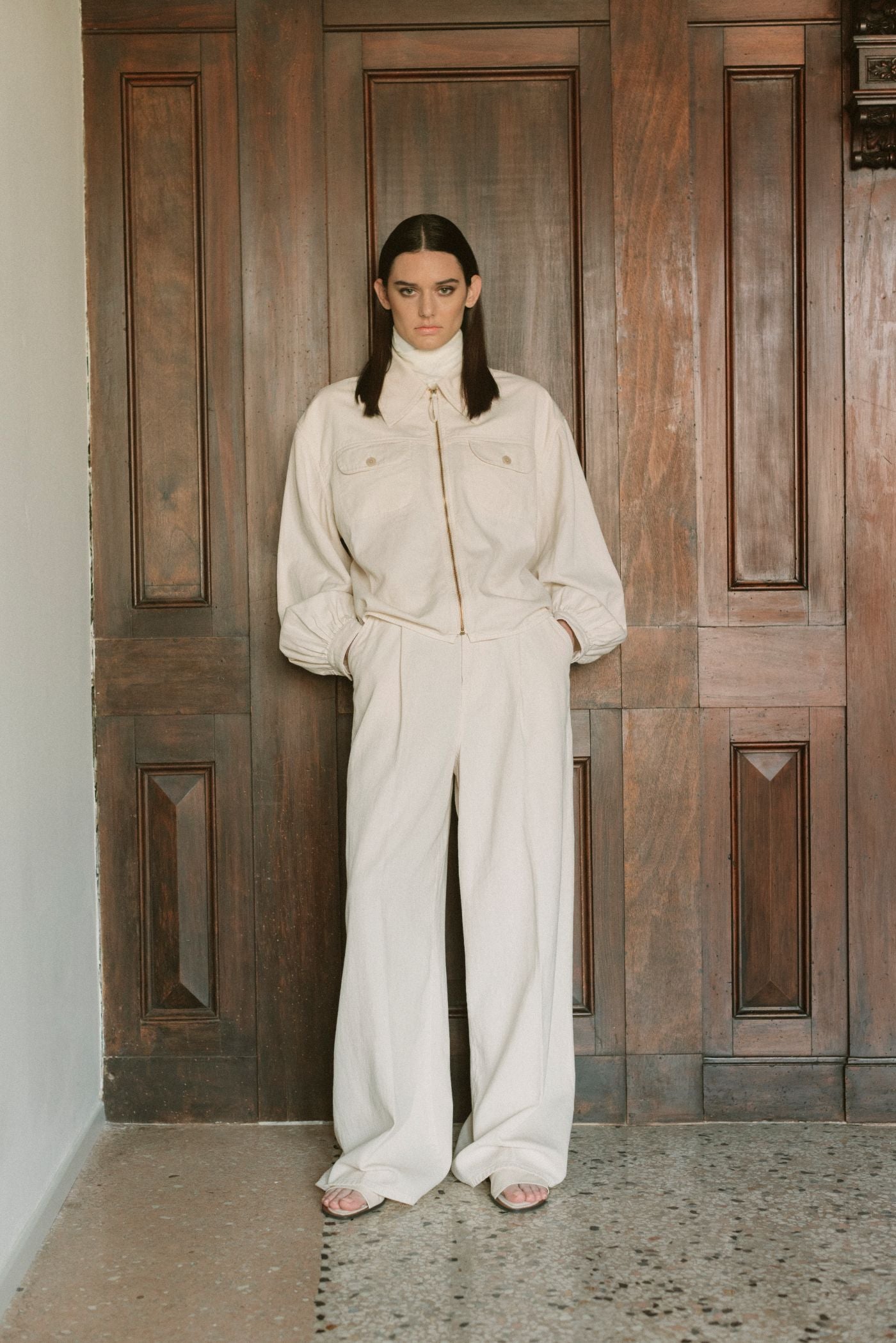 Modella Barmas indossa completo sartoriale chiaro con giacca e pantaloni ampi, in posa davanti a una porta veneziana.