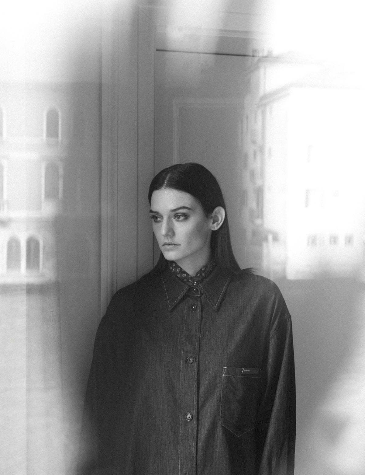 Modella Barmas indossa camicia oversize in denim scuro, ritratta in bianco e nero accanto a una finestra veneziana.