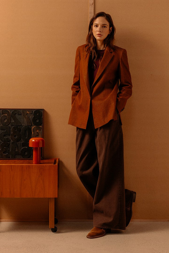 Donna in abbigliamento elegante dai toni caldi con giacca marrone e pantaloni ampi, in un ambiente minimal con mobili in legno e una lampada rossa.