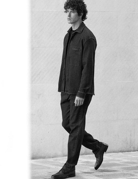 Uomo con capelli ricci che indossa una giacca e pantaloni scuri in stile elegante e sobrio, mentre cammina su una pavimentazione in pietra davanti a un muro chiaro.