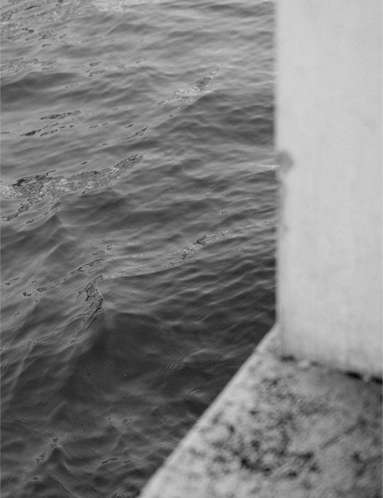 Vista in bianco e nero dell'acqua mossa di un canale veneziano accanto a un muretto di pietra chiara con texture evidente.