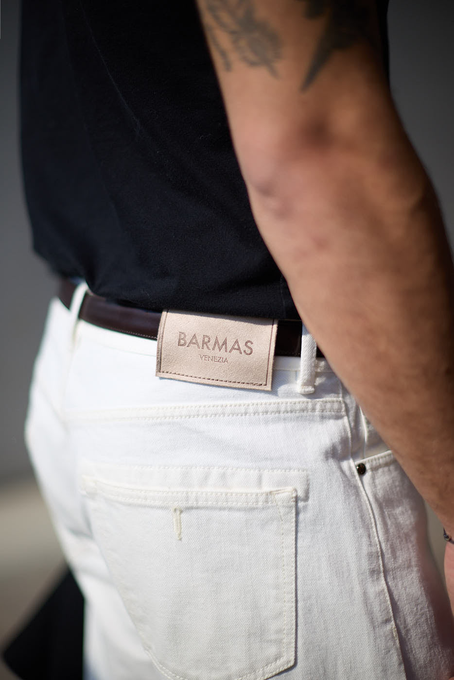 Dettaglio del retro di un paio di pantaloni bianchi in denim con etichetta in pelle Barmas Venezia, indossati da un uomo con cintura marrone.