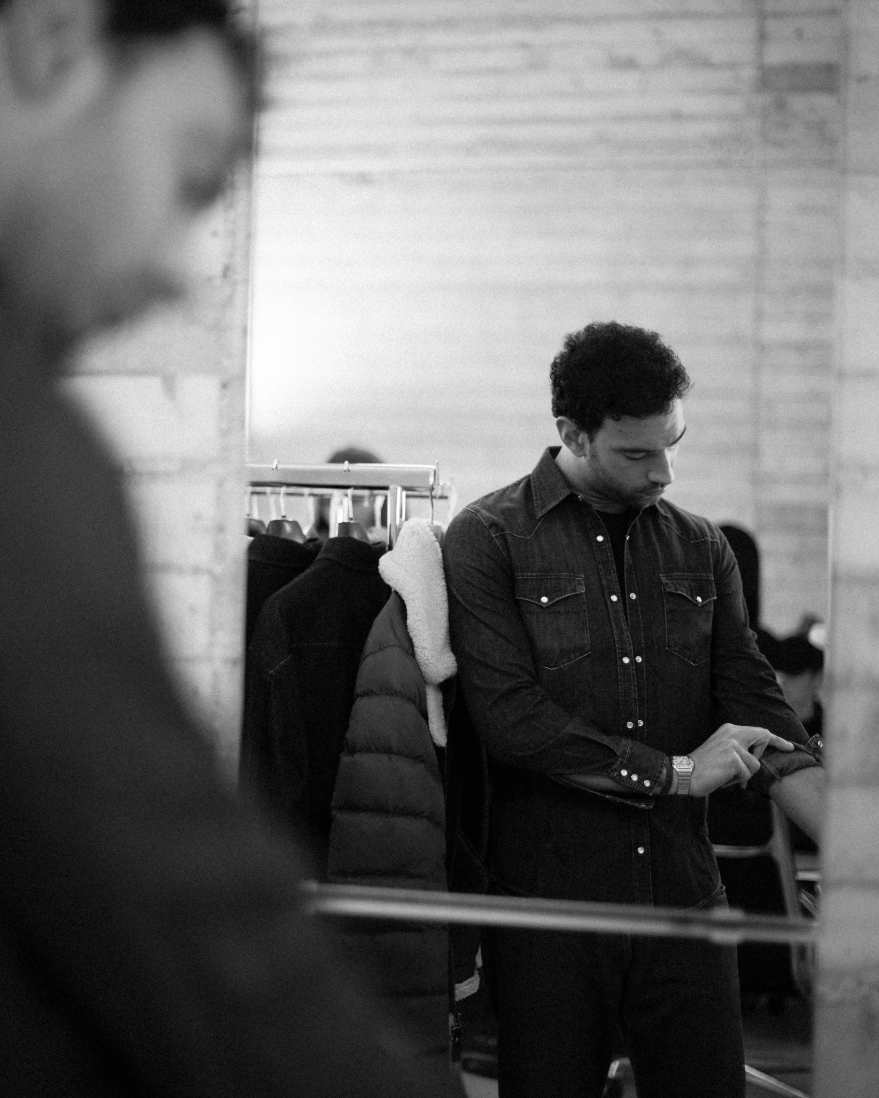 Uomo in camicia di jeans che si guarda allo specchio mentre si rimbocca le maniche, in un atelier con capi appesi sullo sfondo.