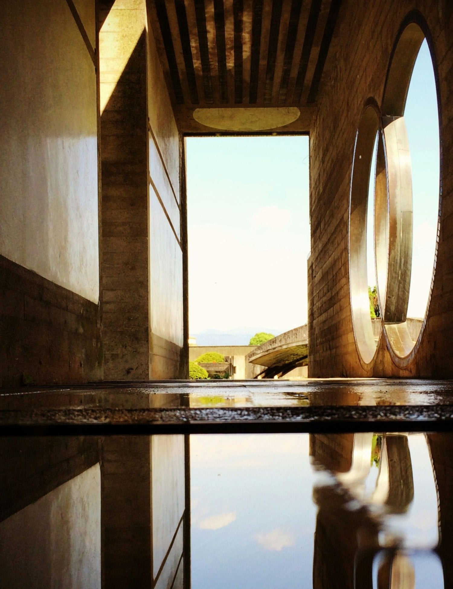 Passaggio architettonico in cemento con ampie aperture circolari, riflesso in una pozza d'acqua che crea un effetto simmetrico; sullo sfondo natura e cielo sereno.
