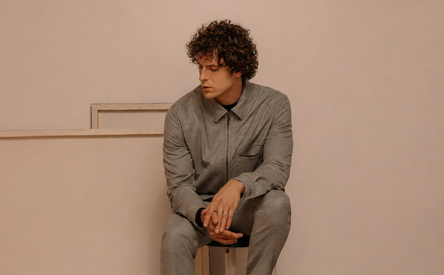 Uomo con capelli ricci castani seduto su uno sgabello in uno spazio dalle tonalità neutre, indossa un completo grigio chiaro dal taglio sartoriale elegante e minimalista.