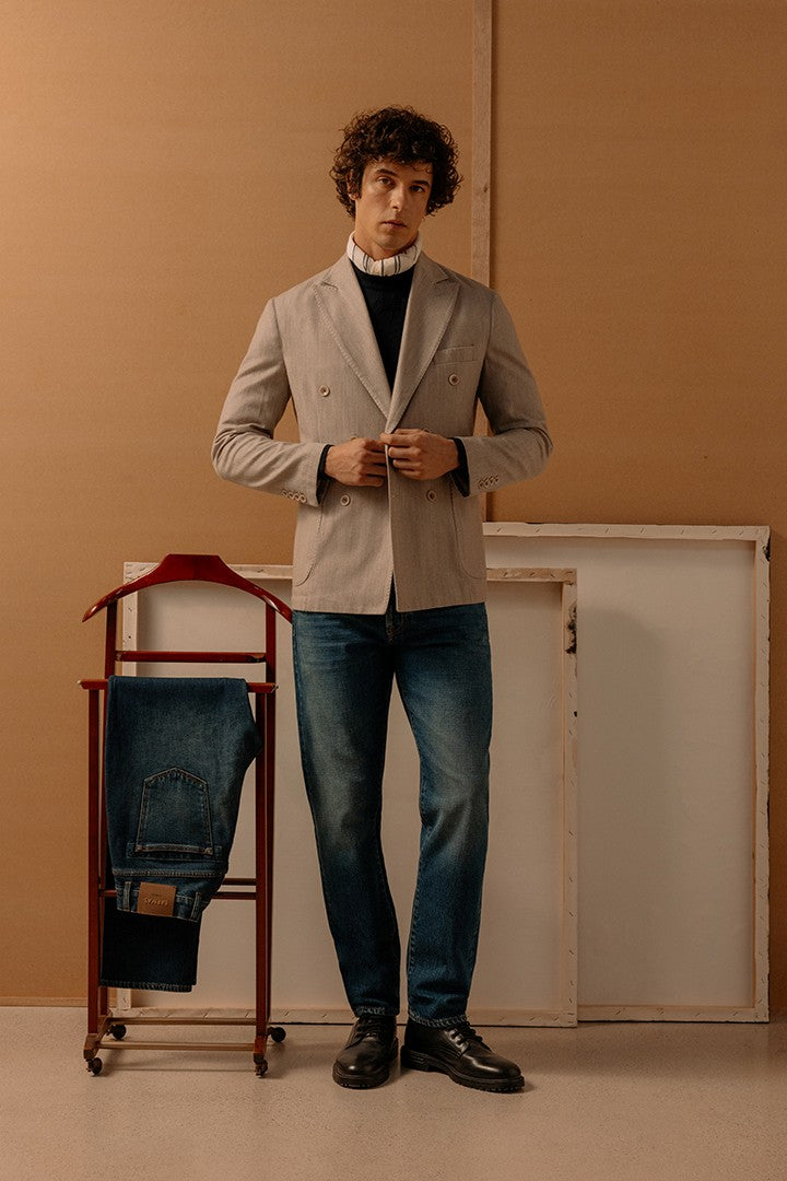 Uomo in posa con giacca beige sartoriale su maglione nero e pantaloni in denim blu scuro, accanto a porta pantaloni con jeans piegati, sfondo neutro e minimalista.