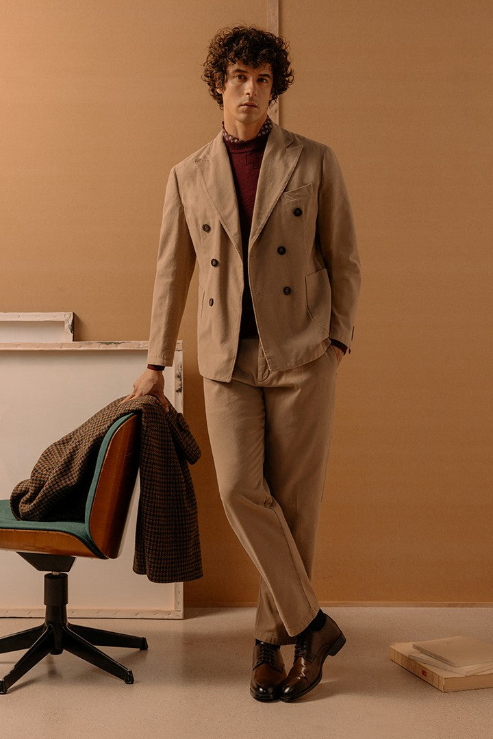 Uomo con capelli ricci indossa completo beige sartoriale con giacca doppiopetto e pantaloni abbinati, maglione bordeaux e scarpe eleganti marroni, ambiente minimalista.