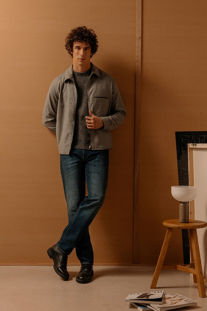 Uomo dai capelli ricci indossa jeans in denim blu scuro e camicia grigia sopra una maglia, posato davanti a una parete marrone con sgabello e riviste a terra, stile sobrio e raffinato.