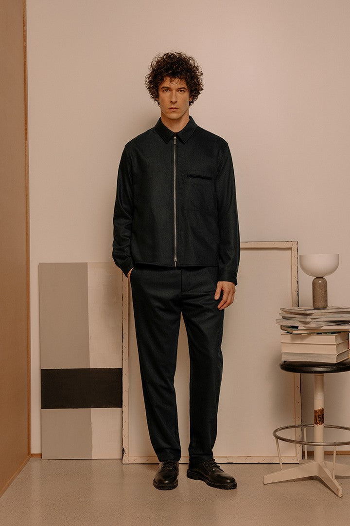 Uomo con capelli ricci indossa completo elegante in tessuto scuro con giacca zip e pantaloni abbinati, in uno stile sobrio e moderno in ambiente minimalista interno.