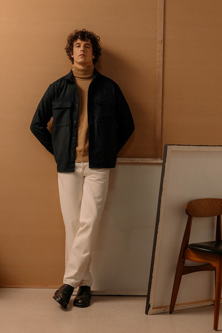 Uomo con capelli ricci indossa un dolcevita color cammello, una giacca nera e pantaloni bianchi, in posa rilassata contro una parete beige con una sedia di legno accanto.