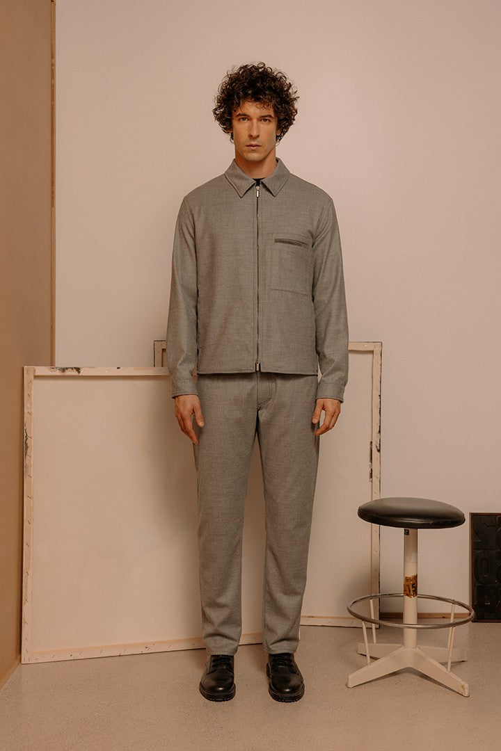 Uomo in piedi con abbigliamento sartoriale grigio composto da giacca con zip e pantaloni, ambientazione minimalista con sgabello e cornici sullo sfondo.