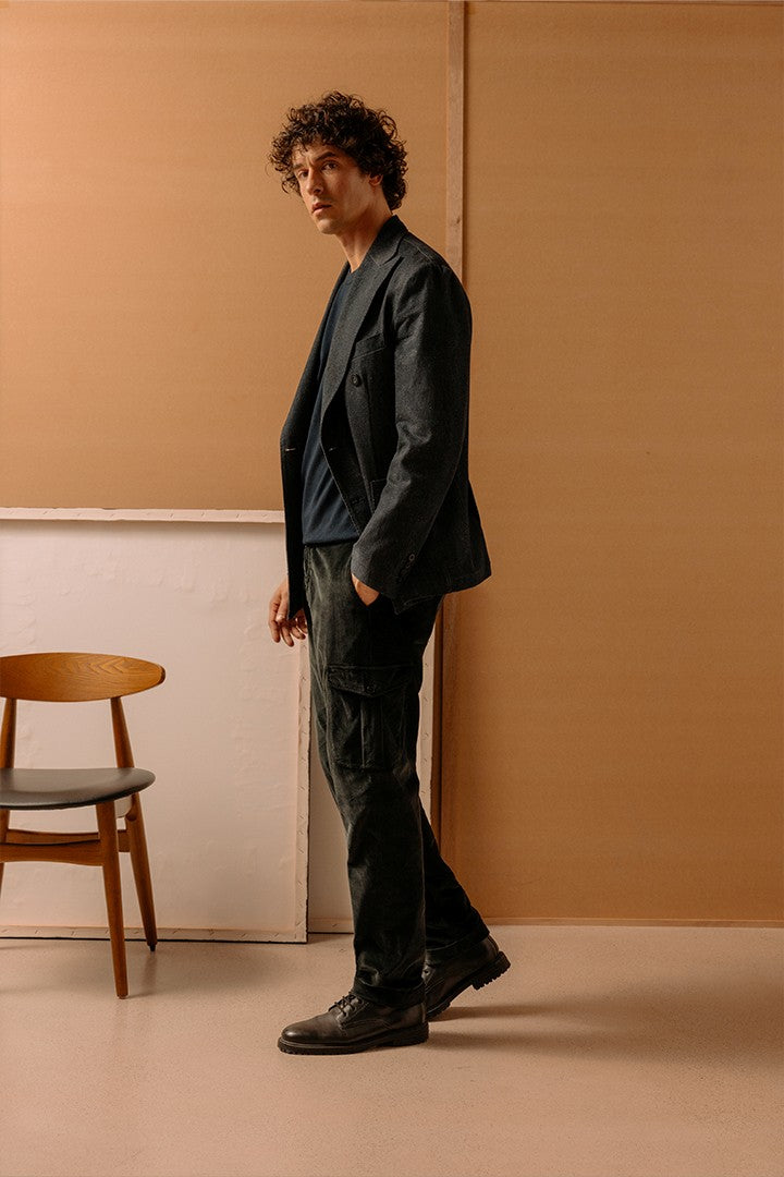 Uomo con capelli ricci indossa giacca nera, maglia blu e pantaloni neri in tessuto tecnico, accostato a una sedia di legno in ambiente minimalista con pareti color beige.